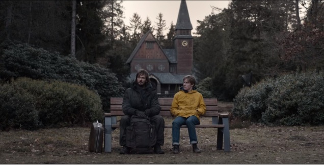 Dwóch mężczyzn, starszy, z plecakiem i walizką, oraz młodszy w żółtej kurtce, siedzą na ławce na cmentarzu, za nimi widać drzewa i kościół,
